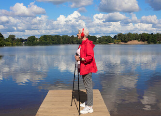 An elderly woman dressed in a red jacket, stands with Nordic walking poles on a bridge by the lake...