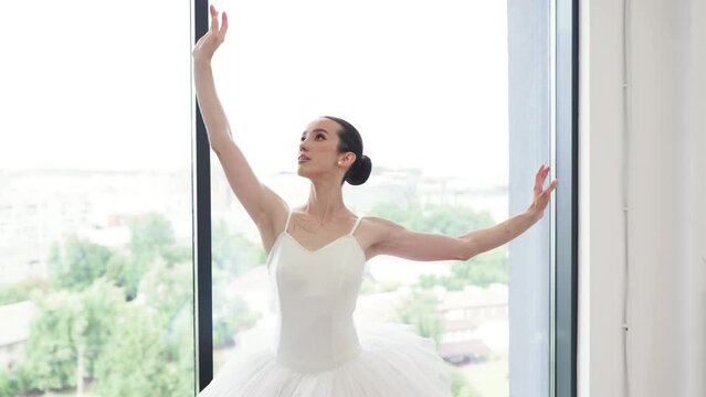 Beautiful graceful ballerina practice ballet positions in tutu skirt near large window in white light hall. Female ballet, contemporary dance teacher class studio. Classical Ballet dancer reverence.