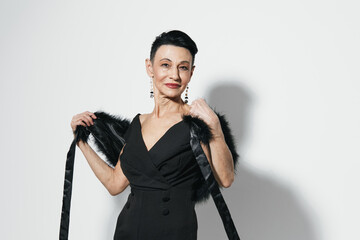 Elegant woman in black dress and fur stole posing confidently against white wall with hands on hips...