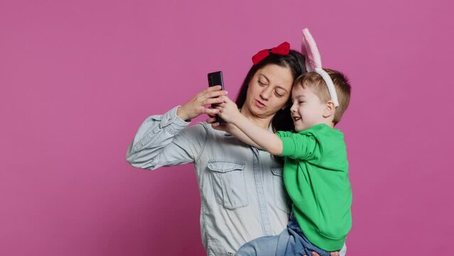 Lovely small child taking pictures with his mother on smartphone, trying to capture fun and cute moments against pink background. Little boy being playful and fooling around with phone. Camera A.