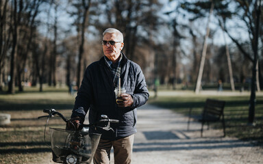 An elderly gentleman strolls through a sunlit park with his bicycle, holding a drink, exuding an...
