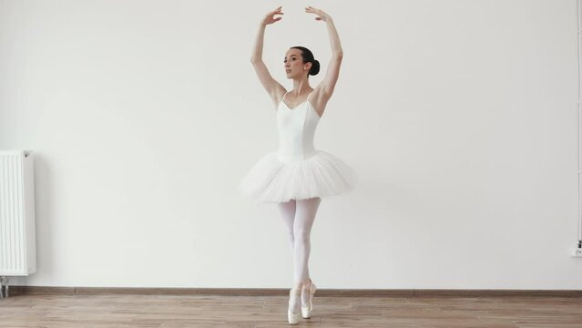 Caucasian young woman ballerina in white tutu, dancing on pointe with arms overhead, in the studio against a light bright background of modern studio hall.