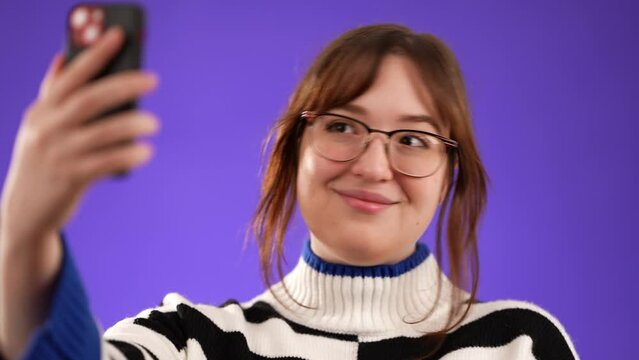 Closeup portrait of happy smiling young woman 20s in sweater taking selfie with mobile cell phone conducting pleasant conversation isolated on purple background studio.