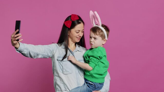 Mother taking photos with her adorable small boy on mobile phone, having fun and kissing each other in studio. Little boy and mom embracing an smiling for pictures on smartphone. Camera A.