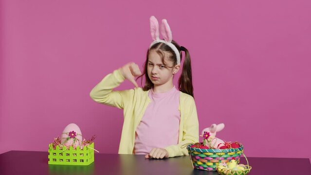 Displeased unhappy child giving thumbs down in studio, expressing her disapproval and disagreement with dislike symbol. Angry dissatisfied young girl showing negative bad gesture. Camera A.