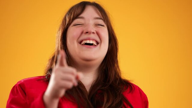 Young woman with brown hair wearing red jacket pointing and laughing, isolated on yellow background with copy space in slow motion