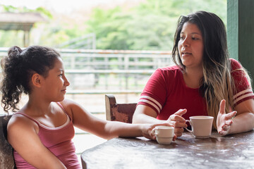 young latina mother talking to her daughter while drinking coffee at the dinner table