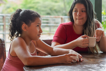 latina brunette girl with a pensive face while her mom watches her and they drink coffee together