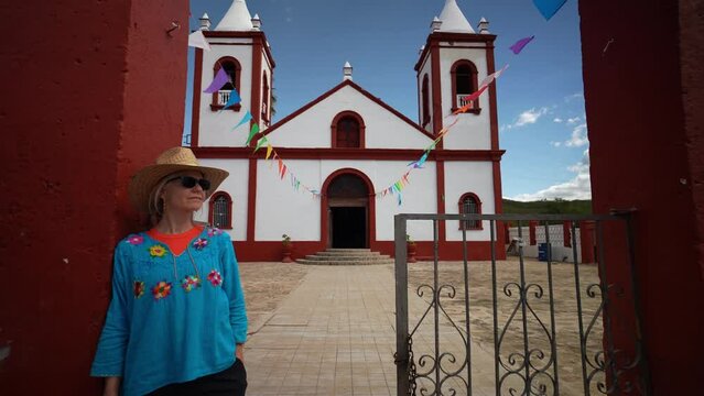 Camera pushes into pretty female tourist wearing straw hat sunglasses, and traditional clothes leaning on wall in front of old church in mining town of El Triunfo, Mexico.