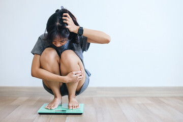Young Asian woman squats on a weigh scale holding her head frustrated over gaining weight.