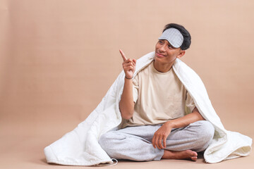 Cheerful young man with sleep mask, sitting on floor covered with blanket while pointing aside with...