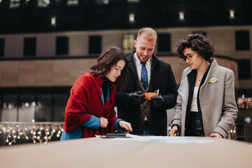 A group of three people engage in a discussion over documents with a smart phone on a city street in the evening.