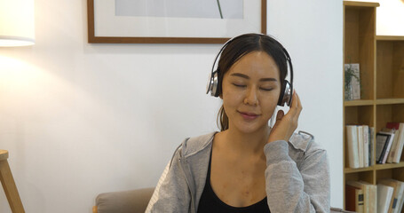 Happy asian woman use headphones listen to music from smartphone outdoor sitting on cozy sofa in...