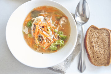 Top view of a healthy soup of meat and vegetables in a white plate on the table with bread and spoon with copy space. Traditional soup with carrots, pieces of meat, broccoli served in plate. 