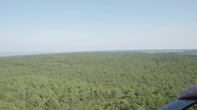 A woman admires the panoramic view of the lush green forest and the sea from the top of the Stilo lighthouse in Stilo, Poland. Pan left shot.