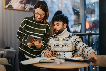 Two young business professionals engage in a collaborative work effort, using a tablet and taking notes in a warmly-lit cafe during the evening.