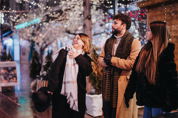 Three young adults are having a bright night out amidst a background of sparkling lights and festive decorations, sharing happiness.