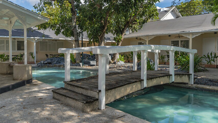 The recreation area. A wooden pedestrian bridge with railings spans over a turquoise swimming pool. The gazebo is nearby. Cottages around the perimeter. Green vegetation. Philippines.