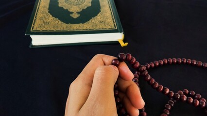 hands holding prayer beads or tasbih, a Muslim is doing dhikr/praying with the background of the Koran or Al Quran