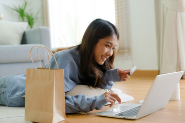Young asian happy woman smile in living room at home, Using computer laptop, lying on the floor at house, online banking payment through the internet from bank card. Shopping online with credit card
