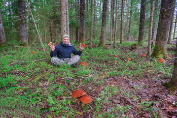 A man sits in a forest clearing and holds mushrooms in his hands. There are many mushrooms growing around him.