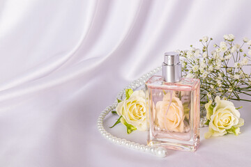 Elegant bottle of women's perfume with delicate floral fragrance on white satin background among tea roses and pearl beads. A copy space.