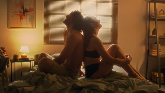 Romantic couple in underwear sitting back to back on bed and daydreaming in cozy bedroom with warm light