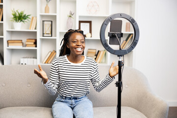 A cheerful Afro American woman engages with her audience on a video call, comfortably sitting at...
