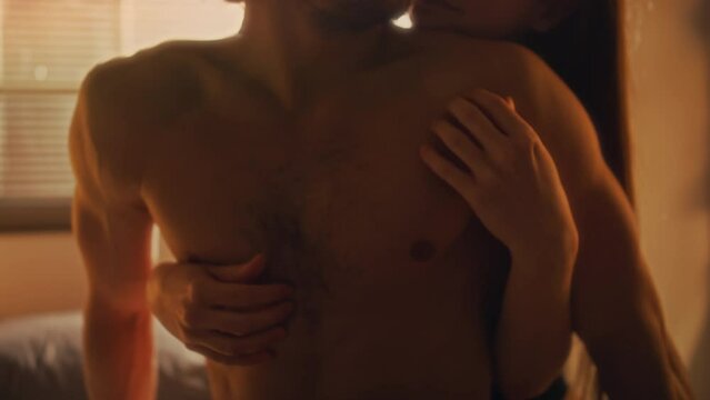 Young woman hugging boyfriend from behind and caressing him in bedroom with warm light