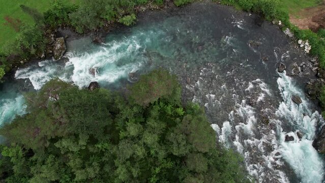 Bird's eye view of a corner in a wide, rapid river. White water is forming as the glacial water crashes over rocks. A dense treeline on the inside of the corner, lush grass and a bench on the outside.
