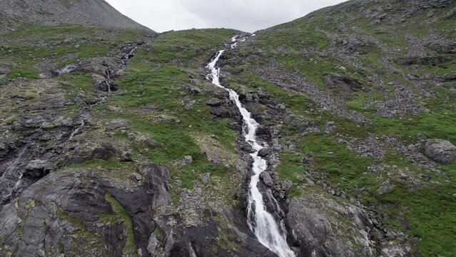Tight aerial dolly shot, pushing in fast on a narrow, rushing river as it winds down a steep, mossy hillside, Trollstigen, Norway. White water cascades down the large rocks along the waters path..