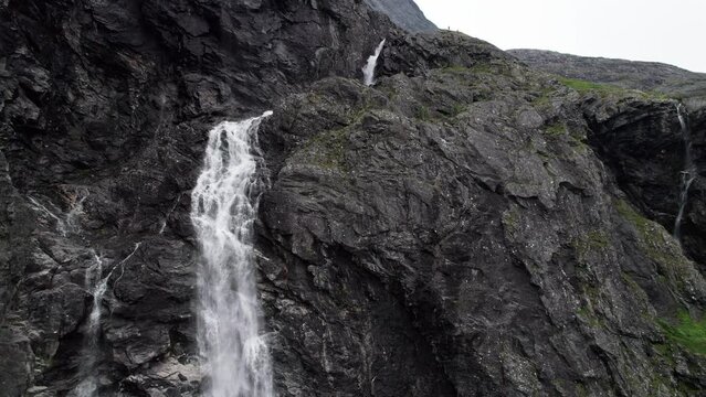 Aerial shot tracking down a cliff, following a waterfall as it cascades down the sheer rock face. Camera pulls back to reveal the whole cliff and the river flows under the Trollstigen road in Norway.