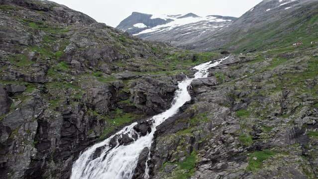 Aerial shot, tracking up from the top of the large Stigfossen waterfall to reveal a large snow capped mountains and a wide valley in the distance.