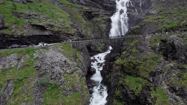 Aerial shot, starting at the Stigfossbrua bridge at the bottom of the Trollstigen in Norway. Camera climbs to reveal the large Stigfossen waterfall crashing down over the sheer rock face.