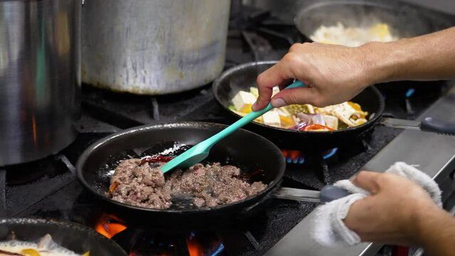Professional restaurant kitchen stovetop filled crowded with multiple occupied skillets cooking a variety of Thai dishes, chef stirs ground beef with red chili peppers, slow motion slider close up 4K