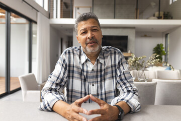 Fototapeta premium Biracial senior man with salt-and-pepper hair sits at a table on a video call, wearing a plaid shirt