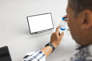A senior man is looking at a tablet with a blank screen, providing copy space, while holding a glass