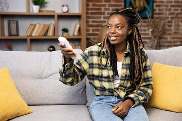 Content African American woman relaxing on couch, utilizing remote control for air con at a...