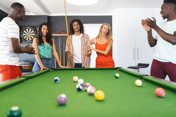 Diverse group of friends enjoys a game of pool, with a young biracial woman taking her shot