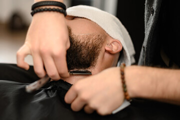 A barber shaves the neck of a bearded customer with dangerous razor. Shaving the contour of the...
