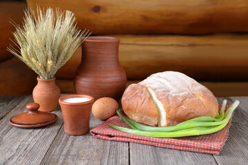 On a wooden background is still life about village food with a loaf of  bread, a clay cup of milk, a hen's egg, green onions, a pottery jug and a vase with ears of wheat. 