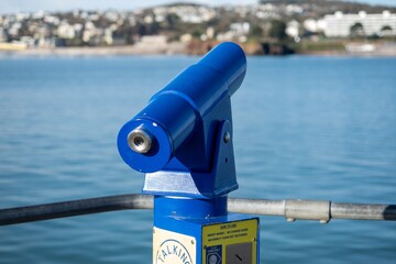 A blue viewing telescope at a seaside location looks across a calm blue sea to land in the distance...