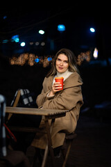 lonely beautiful girl in the evening at a table in a street cafe with a glass of coffee in her hands, night, bokeh
