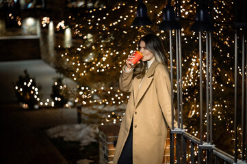 beautiful young girl in a beige coat with a paper glass of coffee against the backdrop of many...