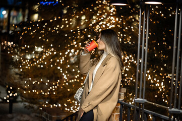 beautiful young girl in a beige coat with a paper glass of coffee against the backdrop of many bokeh lights