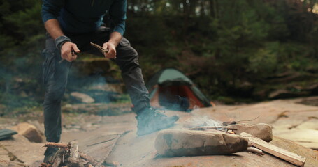 a male tourist chops firewood with a small ax near a campfire in the forest. the traveler prepares...