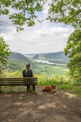 Tourist girl sitting on a bench with a puppy boxer dog looking at the rhine river valley near Andernach from viewpoint - 751186499