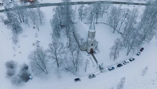 Winter blankets a small church and surrounding cars in a quaint Renda village (Kuldiga parish, Latvia), aerial birdseye orbit view