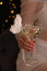 Woman holding glass of cocktail decorated with tasty cotton candy on black background with blurred lights, closeup