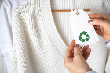 Female hands hold a label from white knitted clothing, which depicts the Recycling sign.
